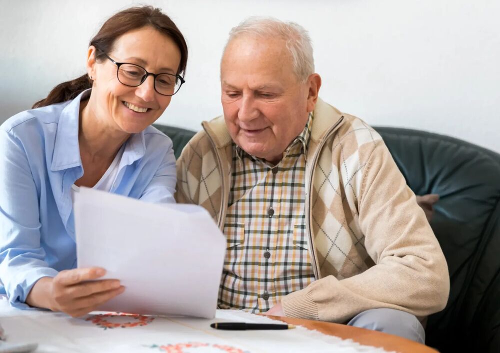Betreuungskraft erklärt einem Senior Unterlagen am Tisch